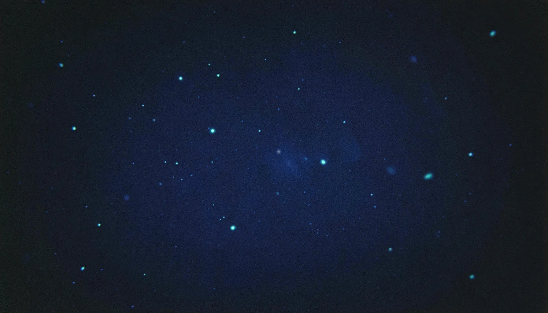 Soft glow of deep cobalt blue with faint luminous cyan particles drifting across a dark navy field, rendered as an analog film photograph with visible grain
